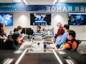 Students sitting around table in the Cyber Range; flat panel monitors hang on the wall behind them